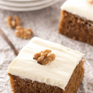 Squares of sweet, spicy pumpkin cake sit ready to eat. The cake is frosted with cream cheese frosting and decorated with walnuts.