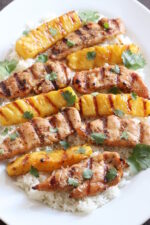 A white platter with a bed of rice. There are strips of grilled chicken breast and strips of grilled pineapple on the rice. The dish is garnished with cilantro leaves.