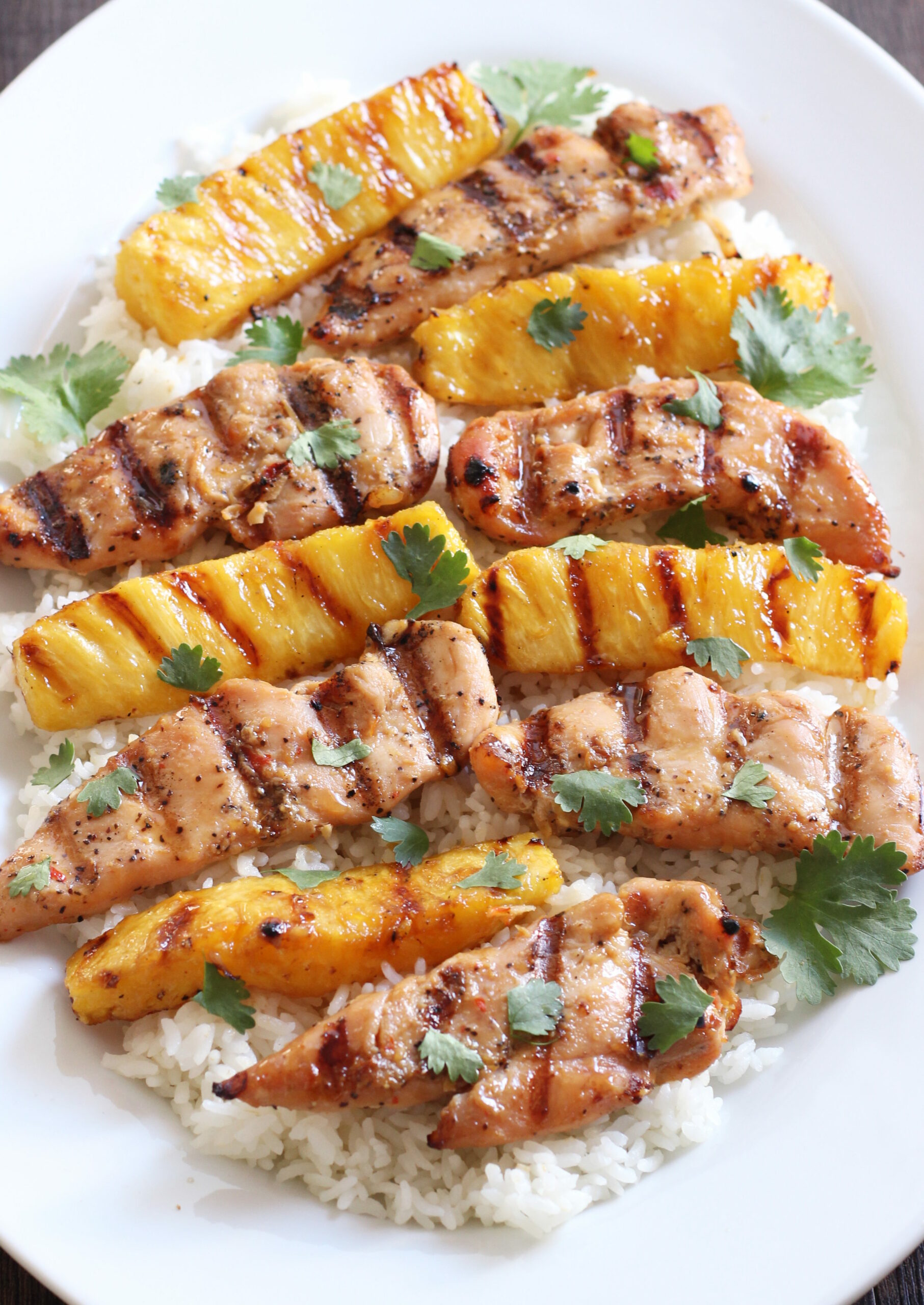 A white platter with a bed of rice. There are strips of grilled chicken breast and strips of grilled pineapple on the rice. The dish is garnished with cilantro leaves.