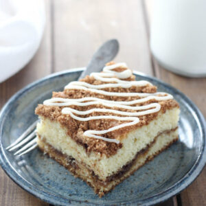 A big square of sour cream coffee cake sits on a blue pottery plate. There is a fork and a glass of milk next to it. The cake has visible layers of cinnamon sugar and a drizzle of vanilla icing.