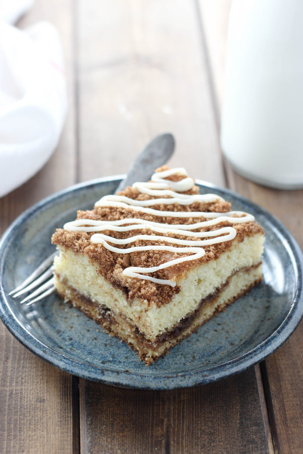 A big square of sour cream coffee cake sits on a blue pottery plate. There is a fork and a glass of milk next to it. The cake has visible layers of cinnamon sugar and a drizzle of vanilla icing.