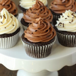 A white cake stand has seven chocolate cupcakes on it. Some cupcakes are frosted with chocolate frosting and some with vanilla buttercream. They all have chocolate sprinkles.