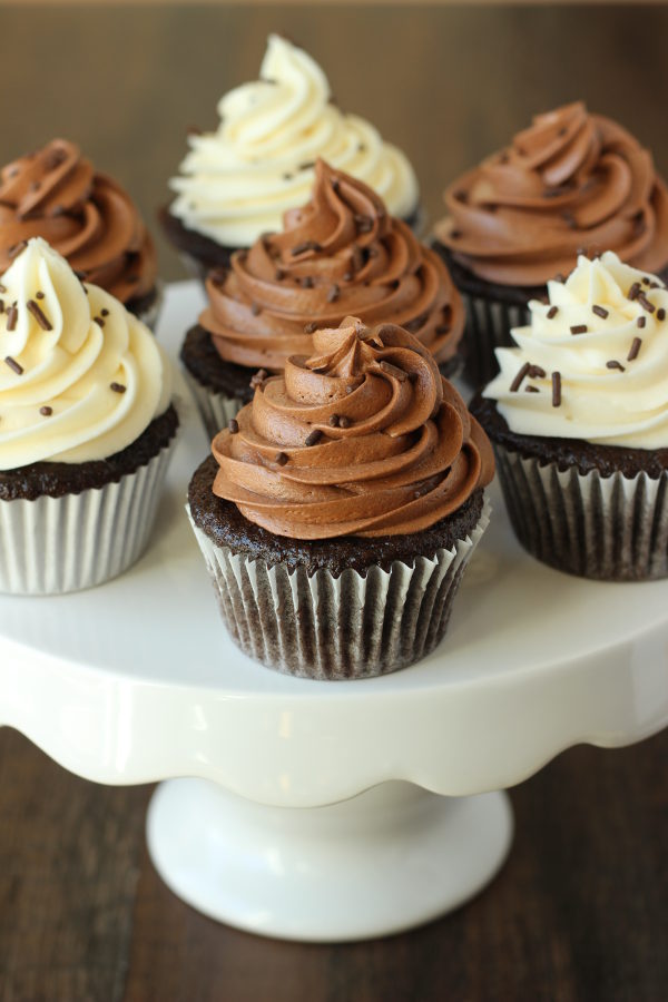 A white cake stand has seven chocolate cupcakes on it. Some cupcakes are frosted with chocolate frosting and some with vanilla buttercream. They all have chocolate sprinkles.