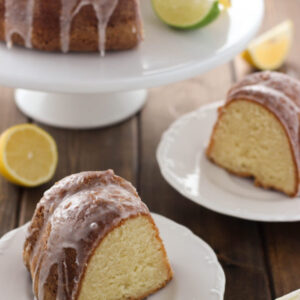 a 70up bundt cake sits on a white cake stand with a slice of lime and a slice of lemon. The cake is glazed with 7 up glaze. Two white plates sit on the table each with a big slice ready to go.