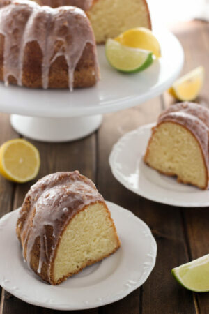 a 70up bundt cake sits on a white cake stand with a slice of lime and a slice of lemon. The cake is glazed with 7 up glaze. Two white plates sit on the table each with a big slice ready to go.