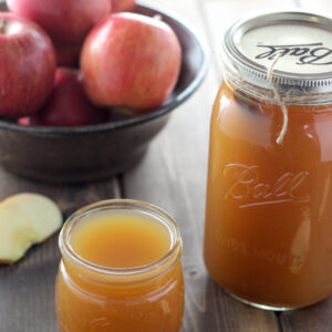 A quart mason jar full of apple pie moonshine sits on a wooden table next to a drinking glass of moonshine and a bowl of apples.