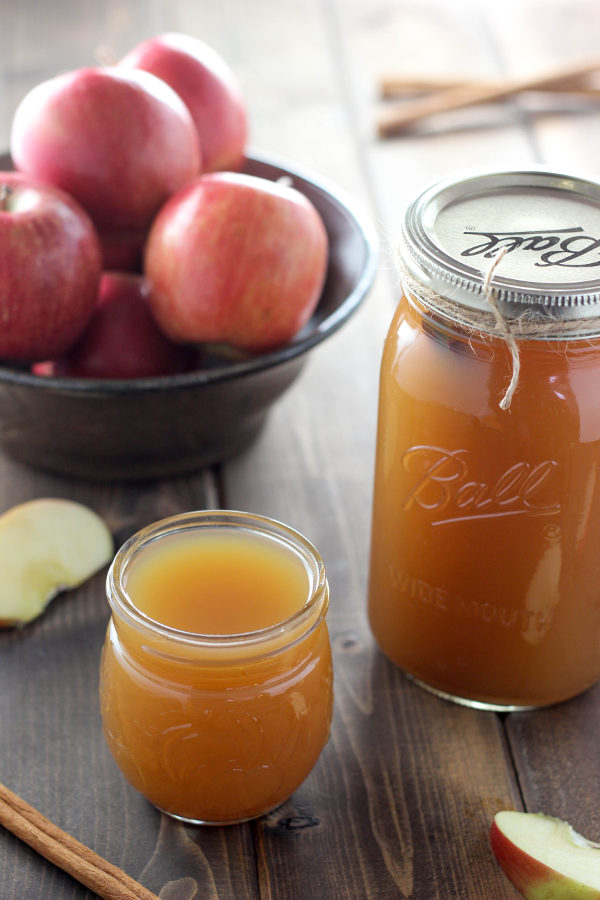 A quart mason jar full of apple pie moonshine sits on a wooden table next to a drinking glass of moonshine and a bowl of apples.