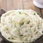 A large serving bowl is full of fluffy mashed potatoes. There is a sprinkling of parsley for garnish and a few pats of butter on the top.