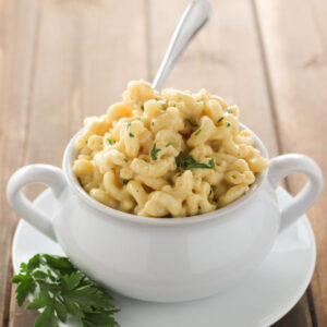 A white bowl is full of saucy, instant pot mac and cheese. The bowl is on a white plate garnished with a sprig of parsley and has a fork sticking out of it.