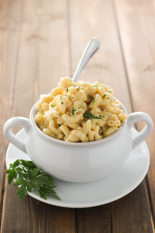 A white bowl is full of saucy, instant pot mac and cheese. The bowl is on a white plate garnished with a sprig of parsley and has a fork sticking out of it.