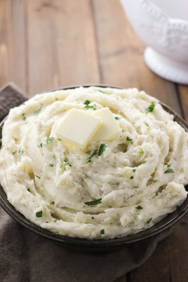 A large serving bowl is full of fluffy mashed potatoes. There is a sprinkling of parsley for garnish and a few pats of butter on the top.