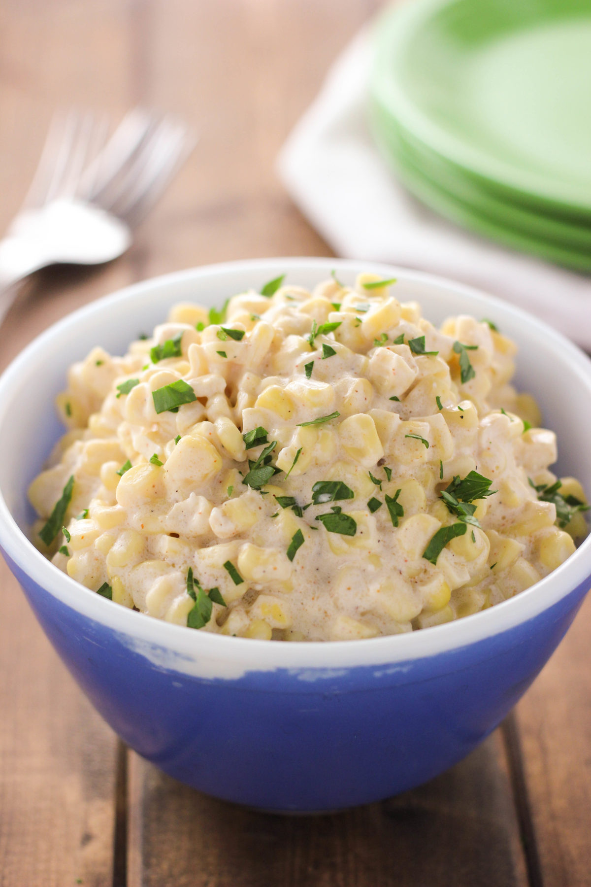 A blue bowl filled with the best creamed corn recipe topped with fresh chopped parsley, shown as a finished slow cooker side dish on a wooden table. You can see a stack of green plates in the background.