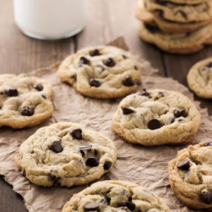 Delicious chocolate chip cookies are arranged on paper on a table. A stack of cookies and glass of milk are in the background.