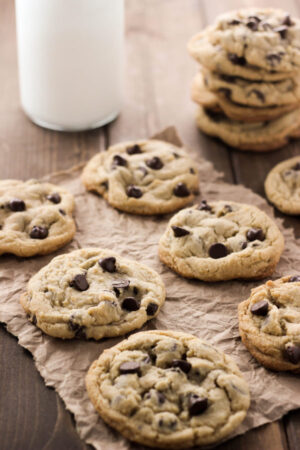 Delicious chocolate chip cookies are arranged on paper on a table. A stack of cookies and glass of milk are in the background.