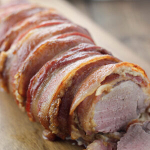 A pork tenderloin is coated in onion sauce and wrapped in bacon. It is sitting on a wooden cutting board getting sliced.
