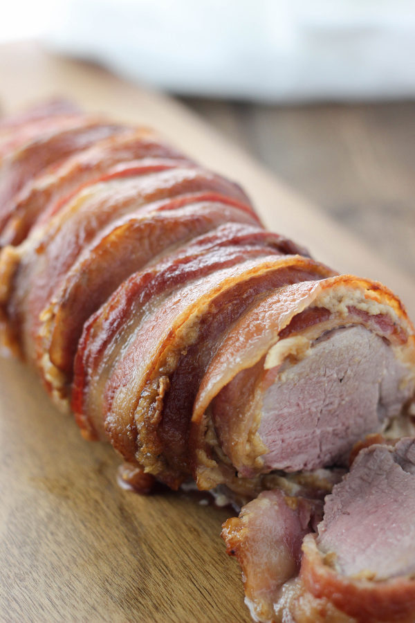 A pork tenderloin is coated in onion sauce and wrapped in bacon. It is sitting on a wooden cutting board getting sliced.