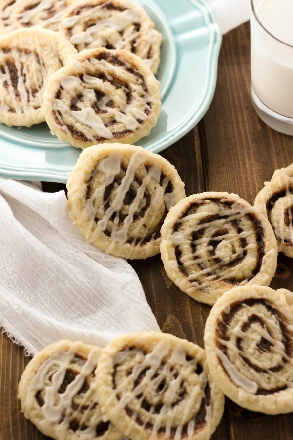 A blue plate covered in cinnamon roll sugar cookies sits on a wooden table. There is a glass of milk at hand to enjoy with the cookies.