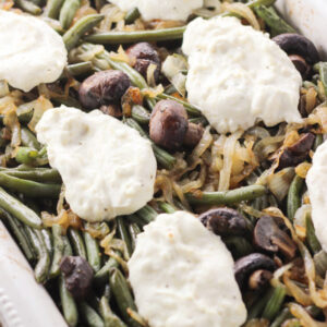 A big rectangular white pan is full of green beans. You can see lots of caramelized onions and roasted mushrooms in this gluten free green bean casserole. There are also big pillows of cheesy, creamy sauce dolloped on top.