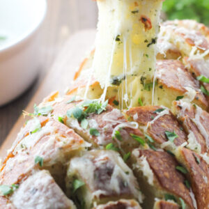 A big loaf of french bread is cross cut and loaded up with butter, herbs and cheese to make this simple cheesy garlic pull apart bread. You can see one piece of bread being pulled out of the loaf.