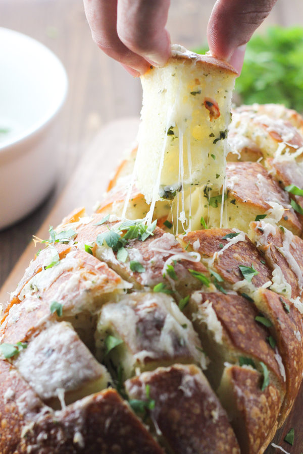 A big loaf of french bread is cross cut and loaded up with butter, herbs and cheese to make this simple cheesy garlic pull apart bread. You can see one piece of bread being pulled out of the loaf.