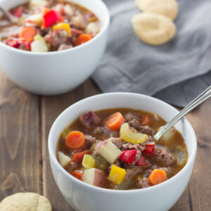 Two hearty bowls of delicious hamburger soup sit on a table with crackers and a gray napkin. You can see big chunks of potato, carrots, and hambuger.