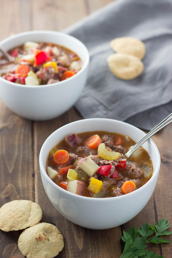 Two hearty bowls of delicious hamburger soup sit on a table with crackers and a gray napkin. You can see big chunks of potato, carrots, and hambuger.