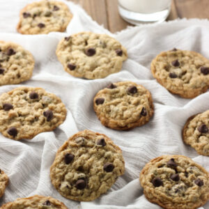 A batch of fresh chewy oatmeal chocolate chip cookies are laid out on a white cloth with a glass of milk.