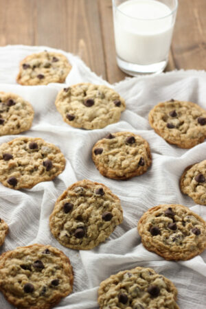 A batch of fresh chewy oatmeal chocolate chip cookies are laid out on a white cloth with a glass of milk.