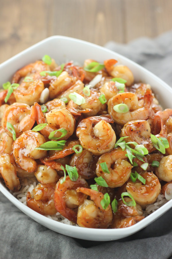 A large white serving bowl is full of shrimp in a golden honey garlic sauce. You can just see some rice under the shrimp and a sprinkling of green onion on top. 