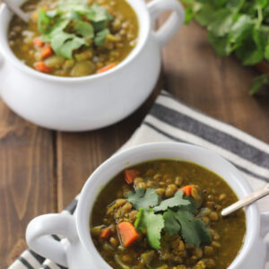 Two white bowls full of curried lentil soup are sitting on a wooden table. Each bowl has mug handles. The soup is garnished with fresh cilantro. You can see carrots and lentils in the yellow-brown lentil soup.