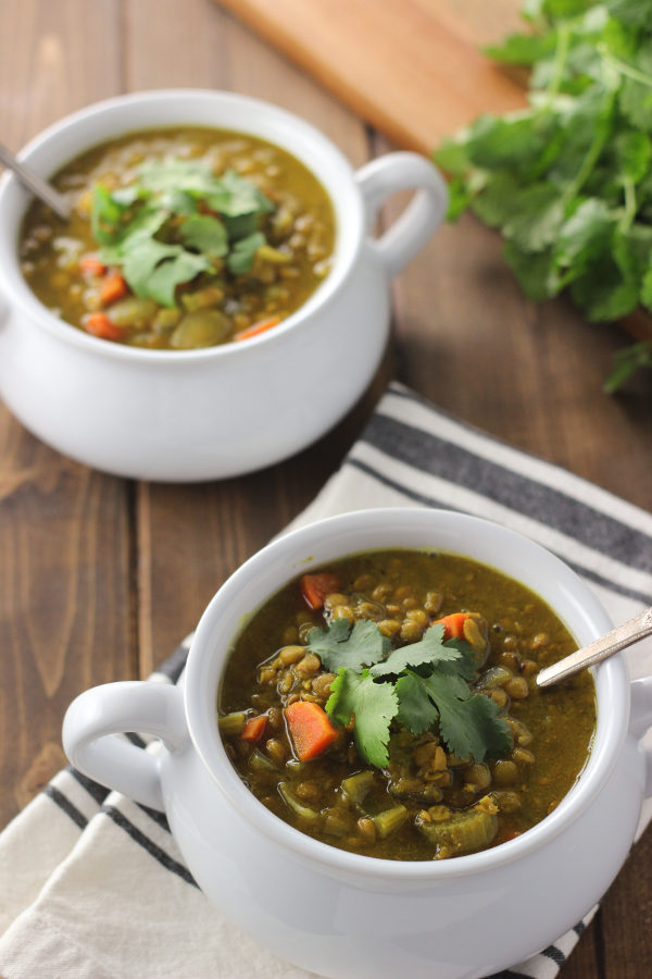 Two white bowls full of curried lentil soup are sitting on a wooden table. Each bowl has mug handles. The soup is garnished with fresh cilantro. You can see carrots and lentils in the yellow-brown lentil soup.