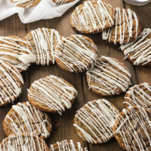 Soft ginger cookies with icing drizzled on them are arranged over a wooden table. There is a glass of milk in the background.