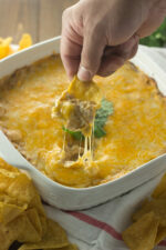 A square white ceramic baking dish is full of cheesy, creamy chicken enchilada dip. A hand holding a chip is in the act of scooping up dip and you can see cheese strings pulling up away from the hot cheese top.