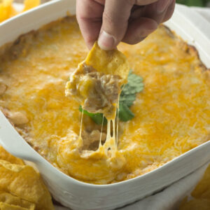 A square white ceramic baking dish is full of cheesy, creamy chicken enchilada dip. A hand holding a chip is in the act of scooping up dip and you can see cheese strings pulling up away from the hot cheese top.