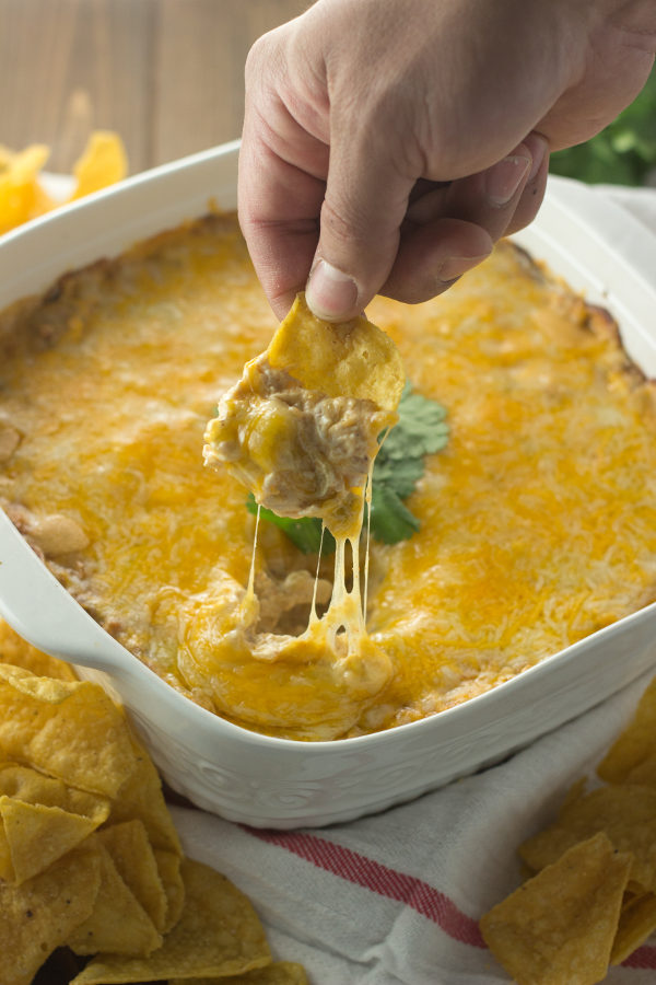 A square white ceramic baking dish is full of cheesy, creamy chicken enchilada dip. A hand holding a chip is in the act of scooping up dip and you can see cheese strings pulling up away from the hot cheese top.