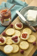 Ritz crackers on a cutting board. You an see a pot of jam and a plate with cream cheese. The crackers have cream cheese, jam and raspberries on them.