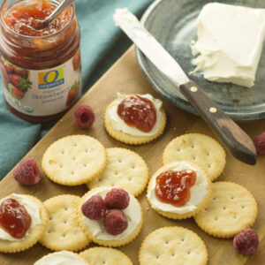 Ritz crackers on a cutting board. You an see a pot of jam and a plate with cream cheese. The crackers have cream cheese, jam and raspberries on them.