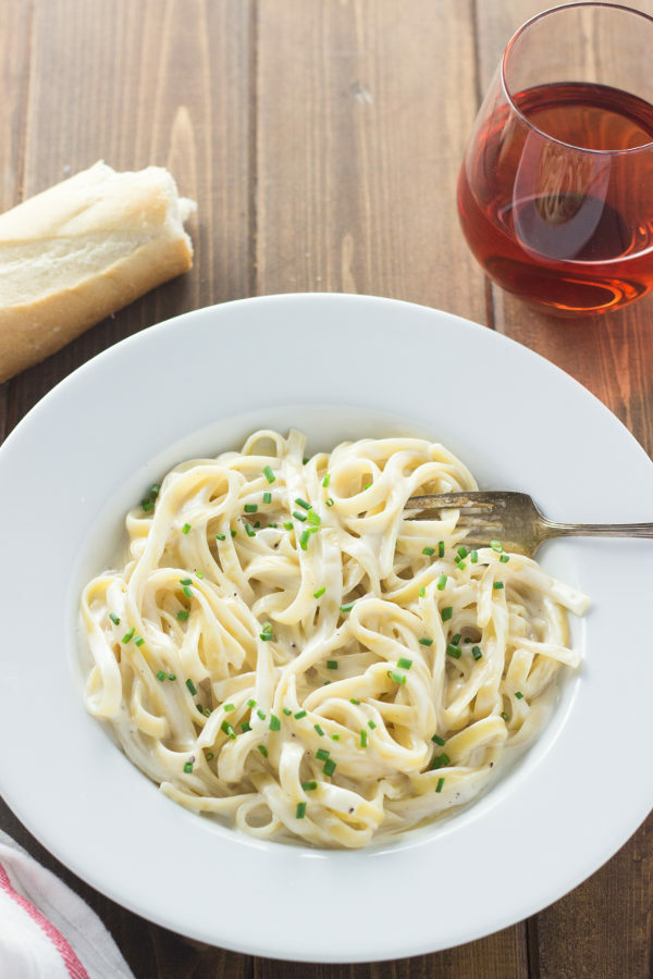 White bowl plate of pasta with 15 minute alfredo sauce from scratch. There is a loaf of french bread on the table and a glass of wine. 