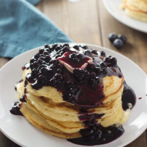 The table is set with two stacks of pancakes. One stack is covered with blueberry syrup and a big pat of butter. The syrup is rich and full of wild blueberries.