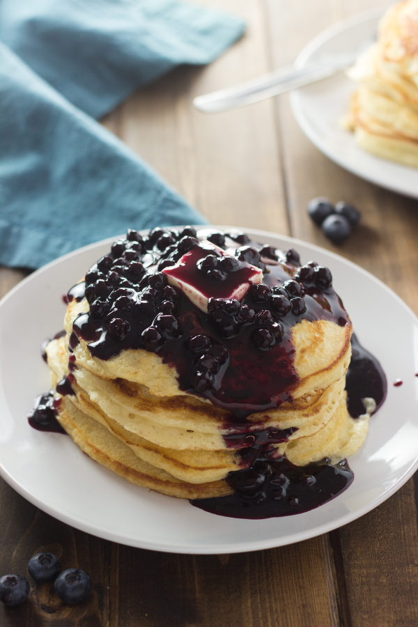 The table is set with two stacks of pancakes. One stack is covered with blueberry syrup and a big pat of butter. The syrup is rich and full of wild blueberries.