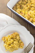 A big square of pineapple casserole sits on a white plate with a fork. You can see the baking dish of pineapple casserole in the background.