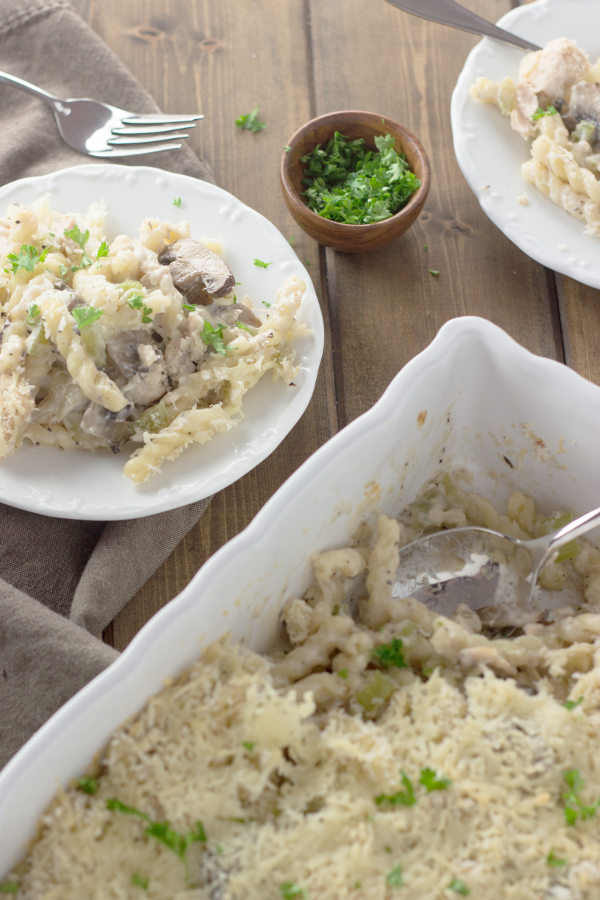 A white casserole dish of tuna noodle casserole is on the table next to a plate of the casserole and some parsley. You can see a few mushrooms and plenty of parmesan cheese in the dish!