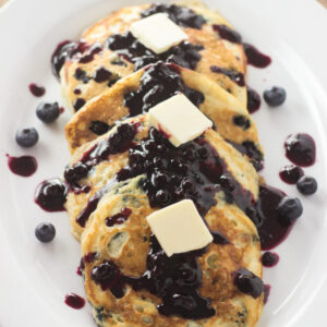 An oval platter is covered in beautiful wild blueberry pancakes. The pancakes have blueberry syrup and pats of butter on them.
