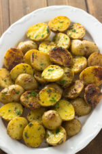 A large white platter of yellow baby potatoes. These potatoes have been sliced in half and oven roasted with garlic and Parmesan. They are finished with butter.