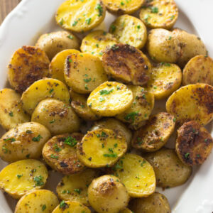 A large white platter of yellow baby potatoes. These potatoes have been sliced in half and oven roasted with garlic and Parmesan. They are finished with butter.