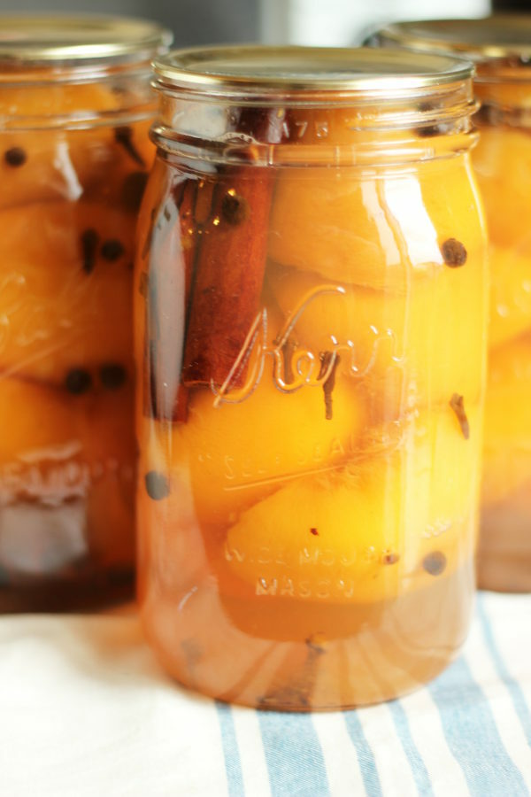 Three mason jars full of peach halves sit on the counter after canning. Each jar is studded with whole spices such as cinnamon, allspice, and cloves. The jars are sitting on a blue and white striped towel to cool. 