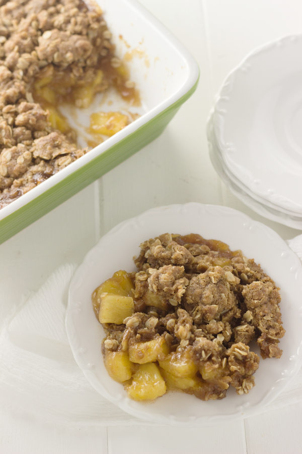 A green casserole dish is full of fresh pineapple crisp. You an see chunks of pineapple where one piece has been lifted out and set on a plate on the table.