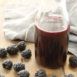 A clear glass bottle of blackberry syrup sits on a wooden table surrounded by fresh blackberries. There is a gray towel in the background.