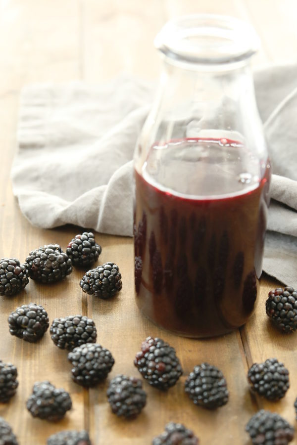 A clear glass bottle of blackberry syrup sits on a wooden table surrounded by fresh blackberries. There is a gray towel in the background.