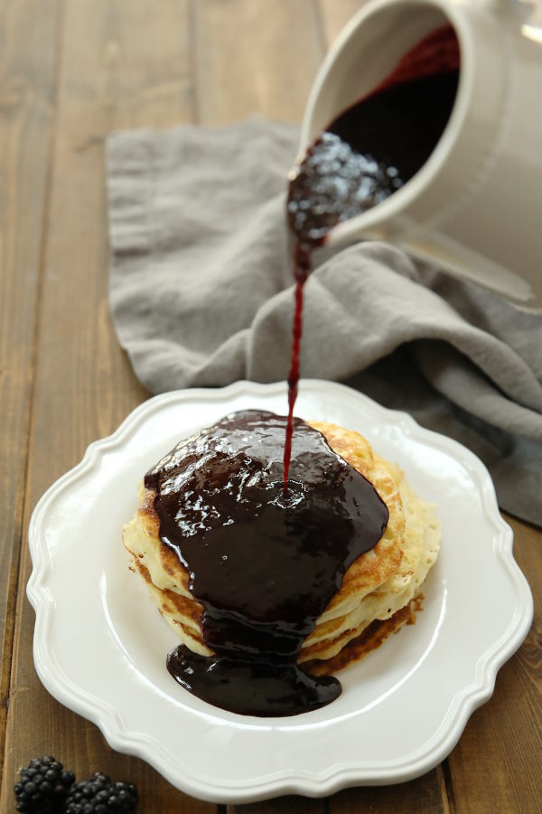 Thick blackberry syrup is being poured from a white pitcher unto a pile of pancakes. The pancakes are stacked on a white plate and there is a gray napkin in the background on the wooden table.